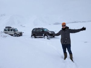 A man in winter clothing smiles in front of two 4x4 vehicles in deep snow with snow-covered mountains in the background in Iceland.