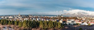 Panoramic view of Reykjavik, with the city, a bay, snow-capped mountains, and evergreen trees under a blue, partly cloudy sky in Iceland.