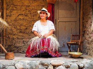 Woman in traditional dress and Panama hat weaving straw outside an adobe home, Ecuador