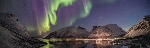 Green and purple aurora borealis shimmer above snow-capped mountains and a town reflected in the water in Iceland.