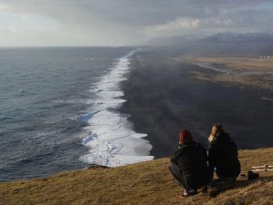 Two people crouch on a grassy hill overlooking a long black sand beach with crashing white waves and distant mountains in Iceland.