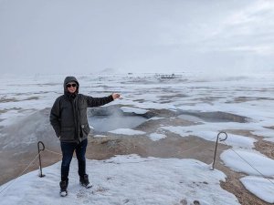 A man in a winter coat stands in a snowy landscape, gesturing toward steaming geothermal pools in Iceland.