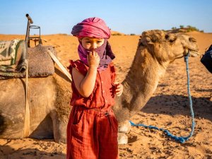 Child wearing face covering in desert next to camel