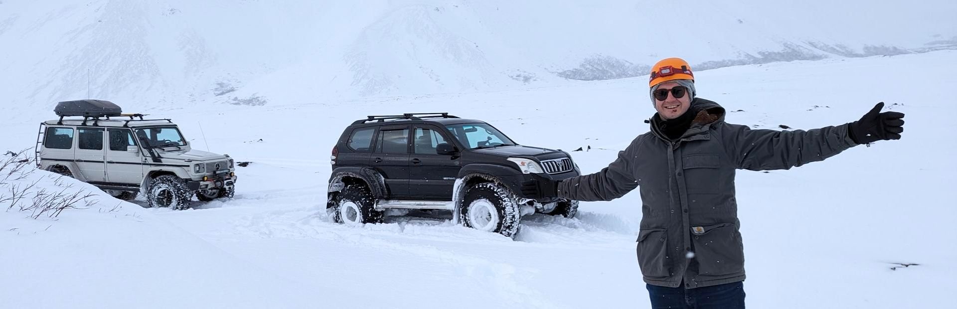 A man in winter clothing smiles in front of two 4x4 vehicles in deep snow with snow-covered mountains in the background in Iceland.