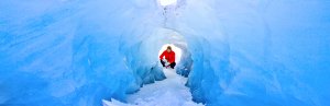 A person in a red jacket crouches inside a glowing blue ice cave in Iceland.