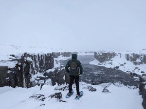 A person wearing snowshoes and a backpack overlooks a river flowing through a snowy winter canyon in Iceland.