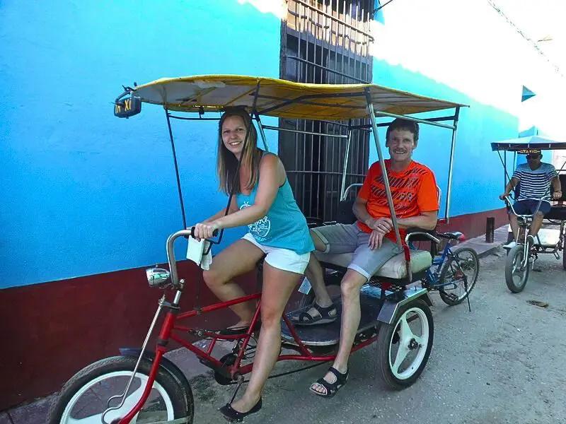 Smiling woman riding a red rickshaw with a passenger in front of a blue wall in Cuba