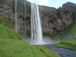 A tall waterfall cascades over a rocky cliff into a stream, surrounded by green grassy hills at Seljalandsfoss in Iceland.