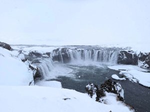 A wide waterfall cascades into a river surrounded by snow-covered cliffs in a winter landscape in Iceland.